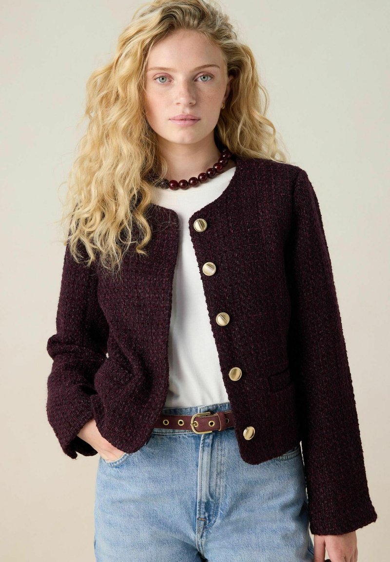 Bordeaux textuurjack met lange mouwen, gouden knopen en een klein zakje. Draagt over een wit shirt en blauwe jeans, met een bruine riem.