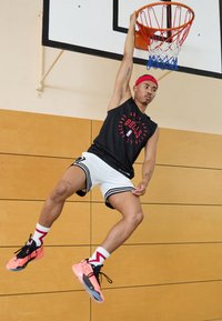 En mandlig atlet hænger fra en basketballring, iført en sort ærmeløs skjorte, hvide shorts med sorte striber og pink sneakers.