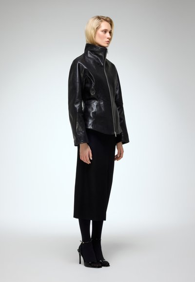 Black leather jacket with high collar and front zipper, paired with a long black skirt and heeled shoes, showcased in a neutral background.