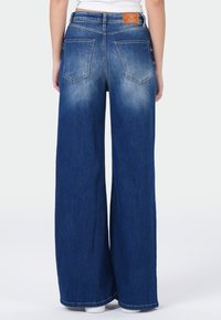 Achteraanzicht van een persoon in brede blauwe spijkerbroek met achterzakken en een leren merklabel op de tailleband.