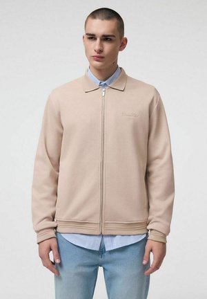 Młody mężczyzna z krótkimi włosami, ubrany w beżową kurtkę na zamek błyskawiczny, nałożoną na jasnoniebieską koszulę na guziki i jasnoniebieskie dżinsy, stojący na jednolitym tle.