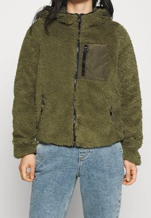 Olivengrøn fleecejakke med lynlås, der har en lynlåslomme på brystet og sidelommer, båret sammen med lyseblå jeans.