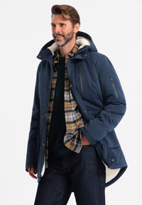 Giacca isolata blu navy con cappuccio foderato in pelliccia sintetica, zip sul petto e sulle maniche, e una camicia a quadri sovrapposta sottostante.
