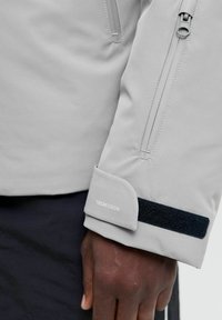 Lys grå jakke med en lynlåslomme på ærmet, blød tekstur, sort velcro-lukning ved håndleddet og "TECHFUSION" logo.