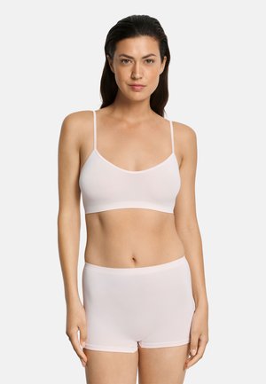 Kobieta w jasnoróżowym bezszwowym biustonoszu typu bralette z cienkimi ramiączkami i dopasowanych bokserkach z wysokim stanem na jednolitym tle.