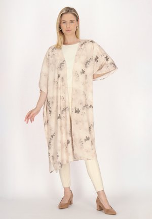 Frau trägt einen hellbeigen Kimono mit Blumenmuster über einem weißen Outfit und braunen Absätzen, steht mit einer Hand auf der Hüfte und neutralem Gesichtsausdruck.