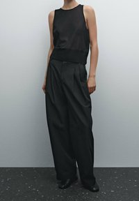 Haut sans manches noir avec une texture transparente, associé à un pantalon noir plissé taille haute et des chaussures noires, sur un fond neutre.