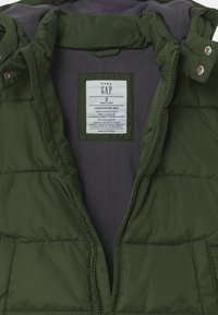 Chaqueta acolchada verde con forro de forro polar gris, tela resistente al agua y una etiqueta que muestra la talla e instrucciones de cuidado.