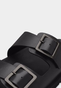 Gros plan d'une sandale noire avec deux larges brides en cuir et des boucles rectangulaires en métal sur un fond uni.