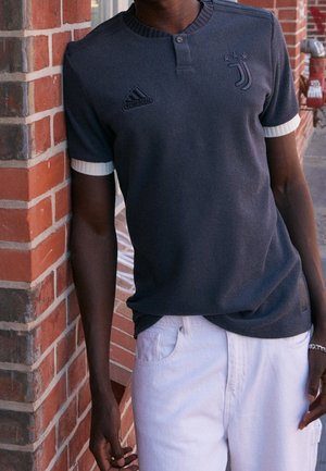 Personne portant un t-shirt Adidas à manches courtes foncé avec le logo de la Juventus et un pantalon blanc, appuyée contre un mur en briques rouges à l'extérieur.