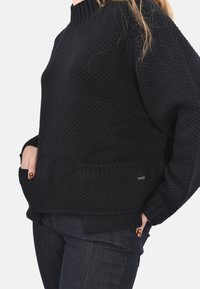 Maglione nero lavorato a maglia con un motivo testurizzato, colletto e polsini a coste; presenta tasche laterali e un orlo posteriore leggermente allungato.