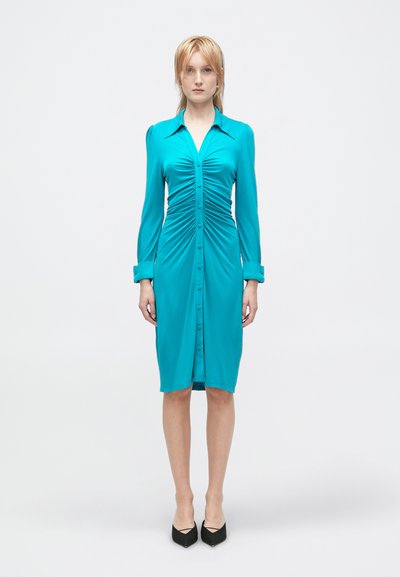 Teal, figurnära klänning med lång ärm, knappstängning fram och samlad struktur längs bodice. Svarta spetsiga klackar kompletterar outfit.