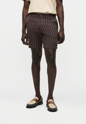 Man wearing dark brown shorts with thin white vertical stripes, beige shirt, and two-tone beige and brown loafers, standing against white background.