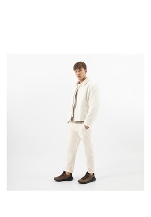 Jeune homme debout portant une veste blanche et un pantalon avec des baskets marron sur un fond blanc, les mains dans les poches, regardant devant lui.