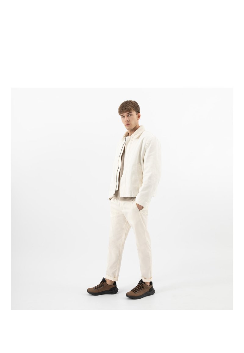 Junger Mann steht, trägt weiße Jacke und Hose mit braunen Turnschuhen, vor weißem Hintergrund, Hände in den Taschen, blickt geradeaus.