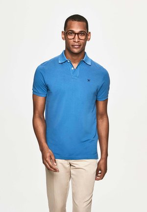 Blaues Poloshirt aus Baumwolle mit kurzen Ärmeln, ausgestattet mit einem Kragen, einem kleinen gestickten Logo und kombiniert mit beigen Hosen.