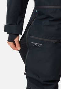 Zwarte outdoor broek met ritsen, voorzien van een mesh binnenvoering, verstelbare manchetten en een logo. Gemaakt van duurzaam, waterafstotend materiaal.