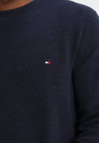 Maglione lavorato a maglia blu navy con texture a coste, caratterizzato da un piccolo logo ricamato rosso, bianco e blu sul lato sinistro del petto.