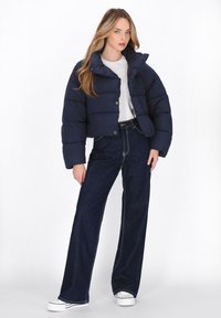Jachetă puffer croppată bleu marin cu guler înalt și închidere cu nasturi, combinată cu blugi întunecați cu talie înaltă și adidași albi.