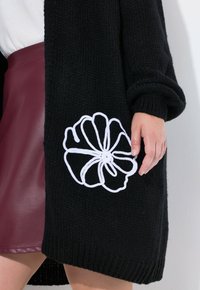 Černý pletený cardigan s bílou květinovou výšivkou na dolní straně. Materiál je silný a má žebrované manžety.