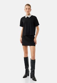 Top negro de manga corta con cuello decorativo blanco, combinado con una minifalda negra y botas de tacón altas negras. Textura de tela suave.