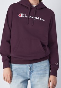Lila hoodie med en framficka, dragsko i huvan och broderad "Champion"-logotyp. Materialet verkar mjukt med en slät textur.