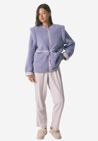 Fioletowa polarowa kurtka z wąskim pasem, szerokim kołnierzem i długimi rękawami. W połączeniu z jasnymi paskowanymi spodniami i beżowymi kapciami.