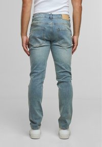 Uomo che indossa jeans in denim blu chiaro e sneakers bianche, in piedi con la schiena rivolta verso la fotocamera su uno sfondo neutro.
