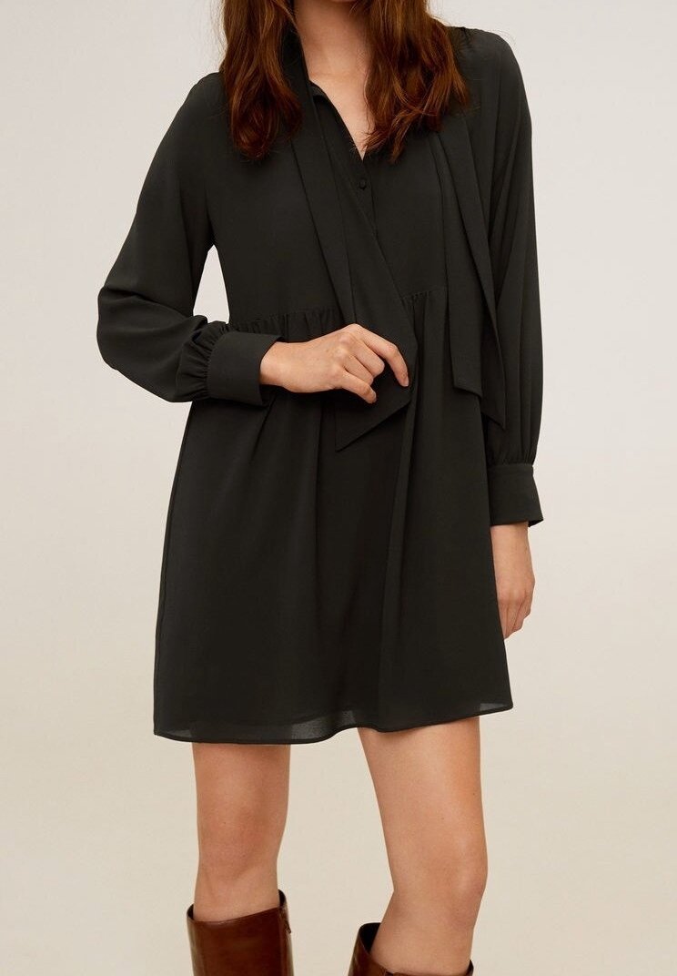 Robe noire à manches longues en tissu léger avec un design plissé à la taille et un col avec des boutons. Accentuée par un drapé doux.