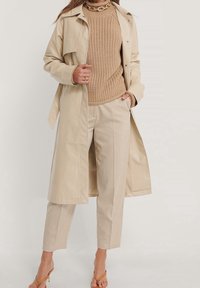 Béžový trenčkot s hladkou texturou, pletený béžový svetr a upravené béžové kalhoty. Celkový vzhled doplňují oranžové sandály na podpatku.