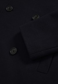 Jūras zila vilnas mēteli detaļas, kas iezīmē dubultā kabatpintes dizainu, gludu tekstūru un melnas pogas ar četru caurumu modeli.