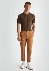 Polo shirt corto a maniche corte in maglia marrone con dettagli a coste verticali, abbinato a pantaloni slim-fit beige e scarpe da ginnastica bianche con suole beige.