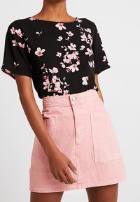 Kvinna iklädd en svart kortärmad topp med rosa blommönster och en hög midja rosa denimkjol med framfickor.