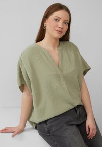 Grüne, strukturierte Bluse mit kurzen Ärmeln und V-Ausschnitt, kombiniert mit dunkelgrauen Jeans. Schlichtes Design, leichter Stoff.