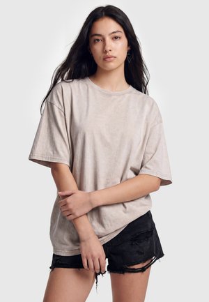 Ung kvinde med langt mørkt hår iført beige oversized t-shirt og sorte revnede shorts, står med korslagte arme mod en ensfarvet baggrund.