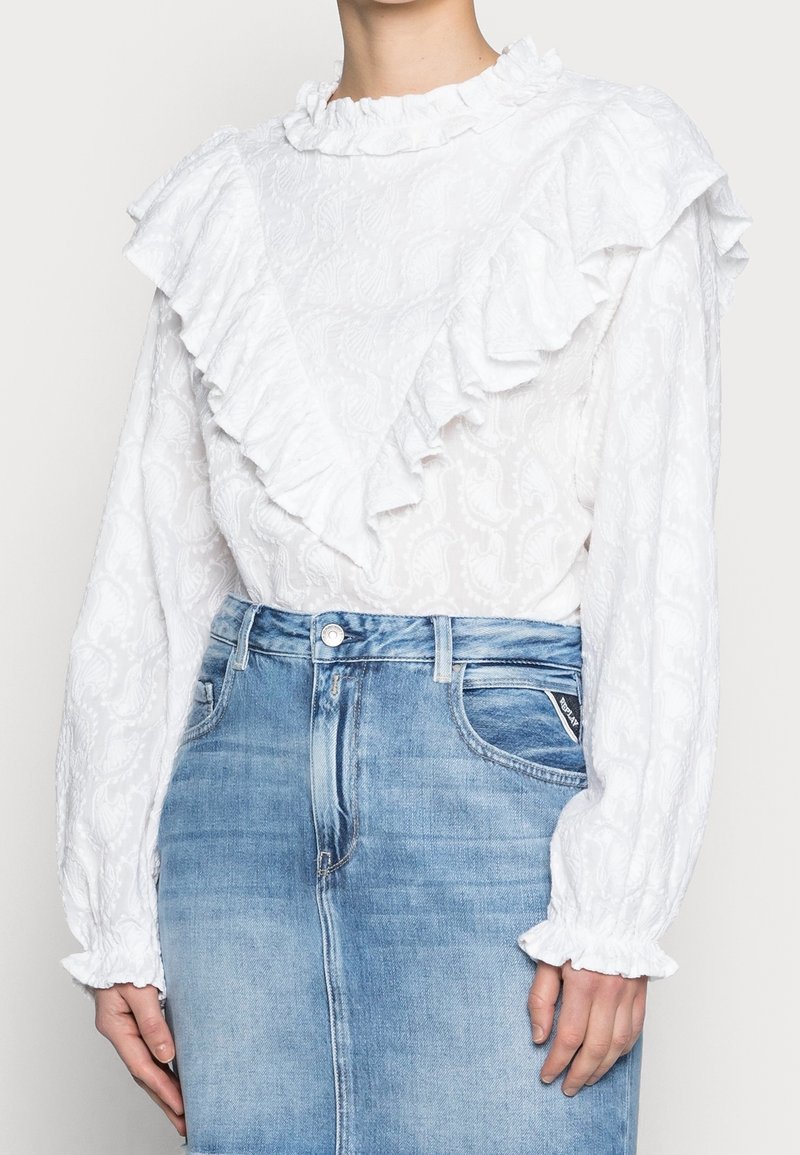 Vit långärmad blus med volangkrage och volanger i ärmsluten, strukturerat blommönster, i kombination med en ljusblå jeanskjol.