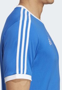 Blå sportt-shirt i bomull med vit tre-stripes-design på ärmarna och en rund vit krage; slät textur.