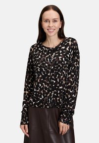 Kamizelka w wzór lamparta w kolorach czerni, brązu i kremu. Długie rękawy, zapięcie na guziki, prążkowana faktura oraz kontrastowe mankiety. Połączona z brązową spódnicą.