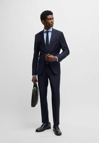 Marineblauer Anzug mit einreihiger Jacke, hellblauem Hemd, schwarzer Krawatte und schwarzen Leder-Schuhen. Trägt eine schwarze Aktentasche.