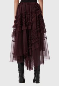 Saia de tule cor burgundy com camadas de babados e um design em cascata, apresentando texturas variadas e uma cintura ajustada. Botas pretas até ao joelho são visíveis.