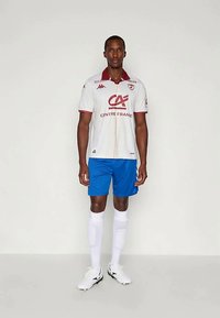 Mužský futbalista stojí v bielom drese s bordovým golierom, modrých šortkách, bielych podkolienkach a bielych kopačkách, pred jednoduchým pozadím.