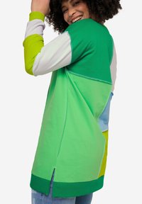 Sudadera de gran tamaño y multicolor con paneles verdes, azules y amarillos, cuello redondo, aberturas laterales y detalles de costura en contraste.
