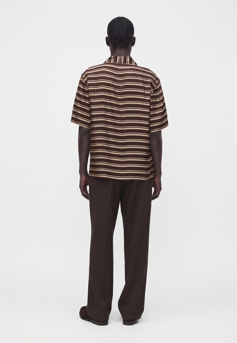Hombre de espaldas que lleva una camisa de manga corta de rayas horizontales marrón y beige y pantalones marrones oscuros, de pie frente a un fondo liso.