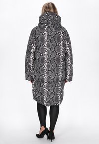 Lang quiltet pufferjakke i sort og hvid slangeskin med en stor hætte, oversized pasform og quiltet struktur; parret med sorte leggings og hæle.