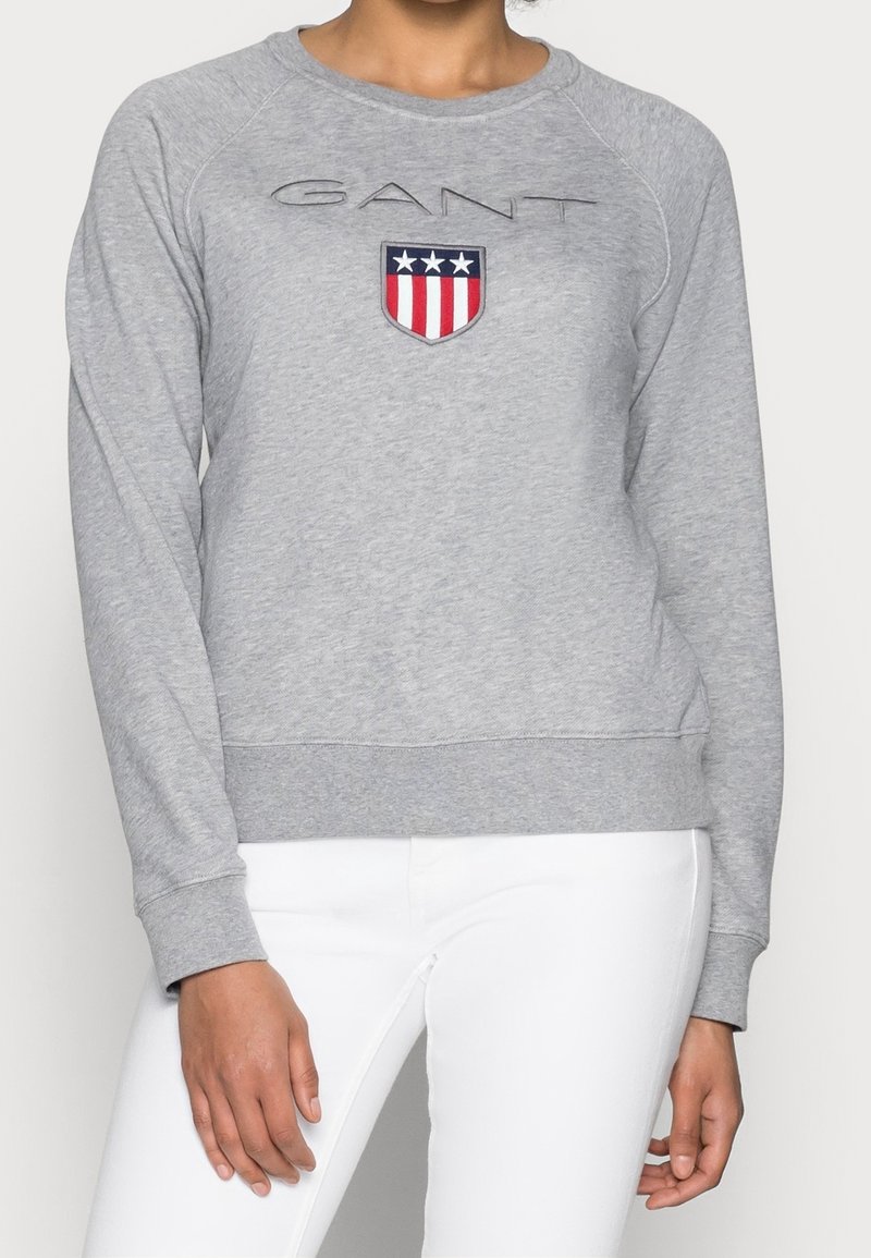Femme portant un sweat-shirt gris clair avec un logo GANT et un design de bouclier rouge-blanc-bleu, associé à un pantalon blanc.