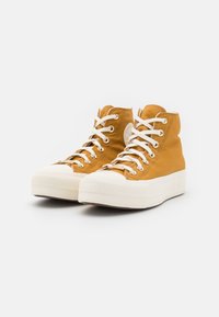 High-top sneakers in mustard yellow canvas with white laces and rubber soles, featuring textured detailing and a logo patch on the side.