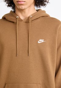 Person iført en brun Nike-hettegenser med hvit logo og snorer, avkortet ved haken og torsoen, mot en ensfarget bakgrunn.