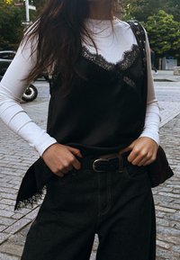 Donna che indossa una maglietta bianca a maniche lunghe sotto un canottiera nera con pizzo, una cintura nera e jeans scuri, in piedi su una strada lastricata.