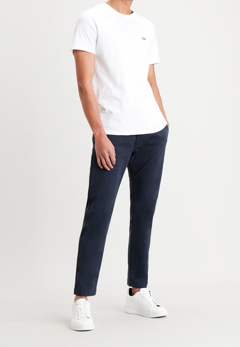 Man in een wit T-shirt met korte mouwen, marineblauwe chinos en witte sneakers, staande met één hand in zijn zak op een effen achtergrond.