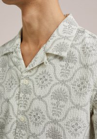 Cămașă cu mânecă scurtă dintr-un material ușor, cu un fundal alb decorat cu modele florale și paisley negre intricate, guler ascuțit și închidere cu nasturi.